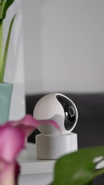 Modern indoor smart home security camera placed on a white side table next to houseplants in a contemporary living room