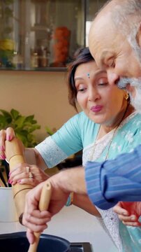 Senior Indian Asian couple enjoying cooking together in their modern kitchen, wife assisting husband in meal preparation, sharing love and joy 