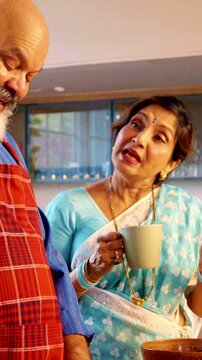 Charming Indian Asian senior couple in a modern kitchen preparing a meal while the husband is cutting vegetables and the wife supervises with a warm cup of tea in hand