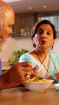 Joyful Indian Asian senior couple enjoying snacks sitting at dining table where the  wife lovingly serves her husband, who enjoys the attention and care