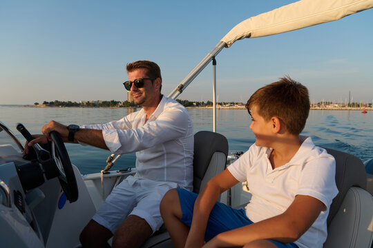 Father And Son Enjoying Summer Boat Ride On Calm Sea Near Coastal Marina During Golden Hour