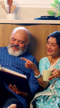 Joyful Indian Asian senior couple sitting comfortably on the kitchen floor, relaxing with cups of tea,fondly looking through an old photo album, reliving cherished memories together
