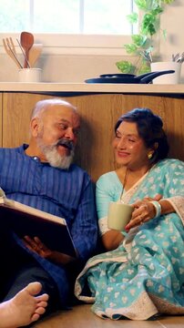 Joyful Indian Asian senior couple sitting comfortably on the floor of their modern, well-equipped kitchen relaxing with  cups of tea, laughing together as they look through an old photo album