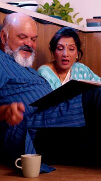 Joyful Indian Asian senior couple relaxing on the kitchen floor sipping tea together, looking through an old photo album and the husband fondly rewinding memories sharing with his wife