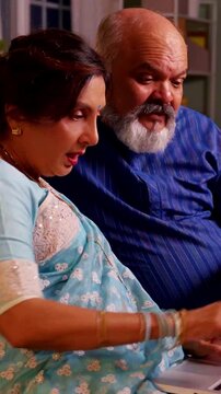 Indian Asian old couple sitting on a sofa at home, working together on a laptop, sharing ideas and enjoying their retired life