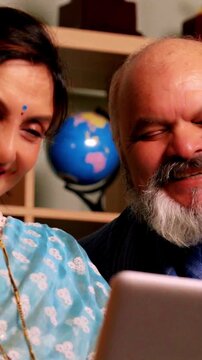 Indian Asian elderly couple sitting on a cozy sofa at home, enjoying a video call with loved ones 