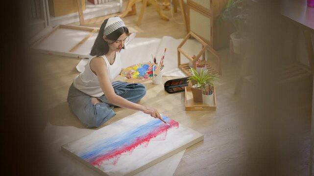 Young Asian female artist sitting on the floor, holding a palette and painting on a canvas framework. Creative girl enjoying painting as an art therapy hobby. lifestyle and home activity.