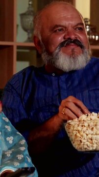 Joyful Indian Asian elderly couple enjoying at home, sharing popcorn while watching their favorite film on television