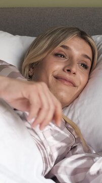 Relaxed young transgender woman in nightwear smiling and rubbing eyes while waking up from bed in bedroom at morning