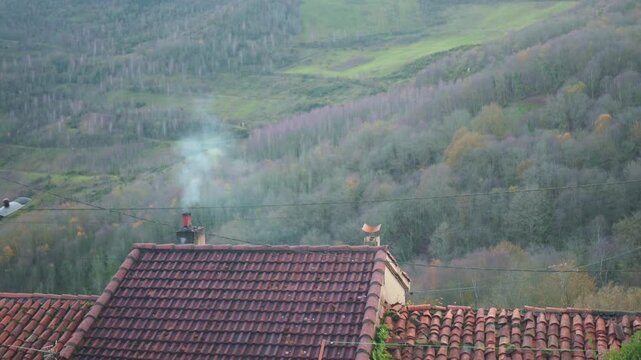 White smoke rising from a chimney on a terracotta tiled roof. A cozy rural house is nestled in a misty valley with bare autumn trees