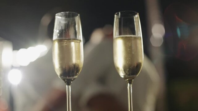 Two elegant champagne flutes filled with bubbly sparkling wine at a wedding reception, set against a blurry, warm, festive background of lights during a nighttime celebration.