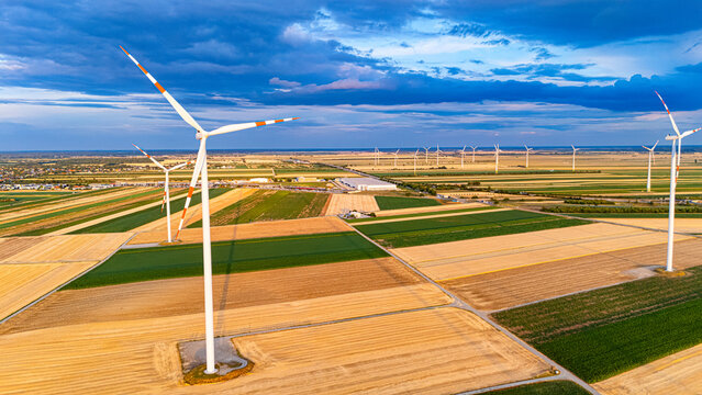 Wind turbines stand tall in the flat landscape during sunrise over fields. Wind turbines spin above vast fields during early morning. The sky has clouds and colors of dawn.