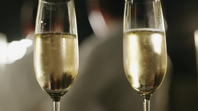 Two elegant champagne flutes filled with bubbly sparkling wine at a wedding reception, set against a blurry, warm, festive background of lights during a nighttime celebration.