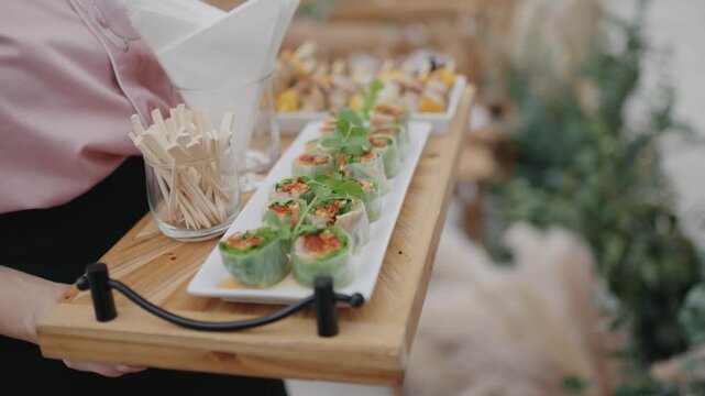 A catering server holding a wooden tray filled with fresh Vietnamese spring rolls and a glass of wooden picks, ready to serve guests at an upscale outdoor wedding event.