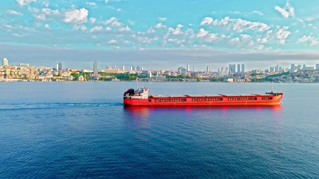 Red cargo ship sailing past Dolmabahce Palace on the Bosphorus in Istanbul, Turkey. Calm blue water, soft clouds and the palace shoreline create a scenic maritime view.