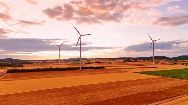 Wind turbines generating power in golden fields at dusk. High angle view of three alternative energy structures under orange clouds and pink sky during evening sunset.