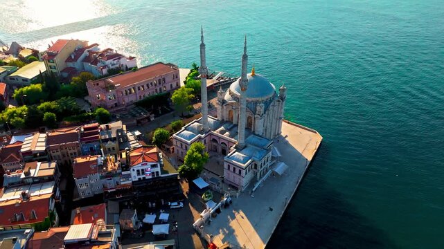 Drone view above Buyuk Mecidiye Mosque and the surrounding Ortakoy waterfront. Calm blue water and the Bosphorus shoreline frame this scenic Istanbul landmark.