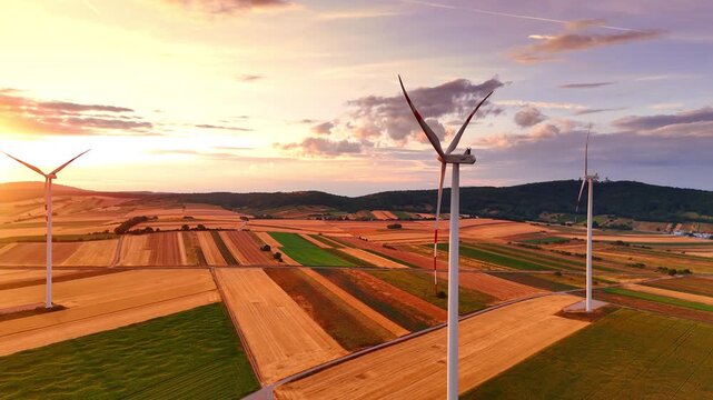 Wind turbines spinning over agricultural landscape during warm sunset. Drone perspective of ecological power generators among orange fields with dark green mountains on background.