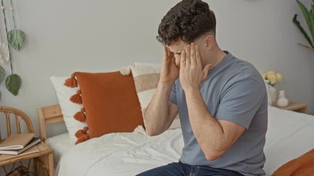 Man rubbing temples with hands on bed beside a tasseled pillow in building, touching face for headache and visible discomfort; fatigue.