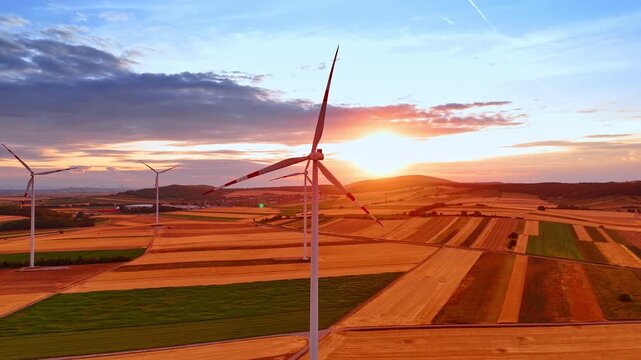 White wind turbine silhouette against bright orange sunset. Aerial view of eco power generators on rolling fields during golden hour light.