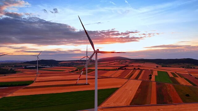 Industrial wind turbine generator standing in golden sunset. Wide aerial viewpoint of modern eco technology over striped agriculture field landscape.