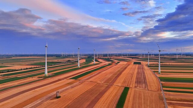 Wind farm turbines standing on colorful agricultural fields. High angle drone shot of alternative energy generators under pastel sunset clouds.
