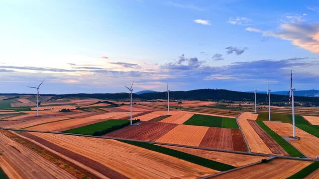 Aerial view of wind turbines across patterned countryside. High angle drone shot of clean power station on striped green and yellow fields.