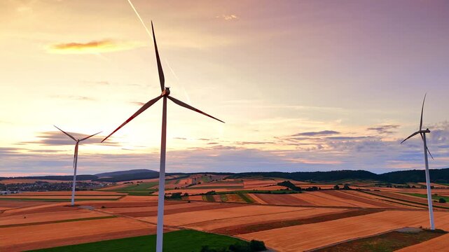 Wind turbines over agricultural fields during vibrant sunset. Aerial view of three wind generators on rolling farmland under orange and purple sky.