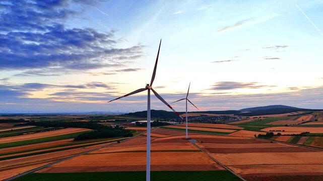 Alternative energy turbine operating in rural farmland landscape. Wide drone view of a clean power system over striped fields under evening sky.