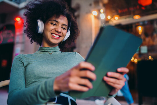 young student watching streaming video content on a wireless tablet device display while using white headphones inside street cafe, happy Gen Z youth browsing social media network app
