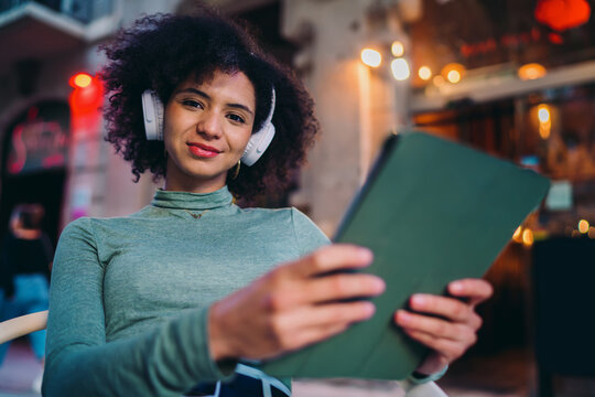 young user checking dynamic notifications feed on modular touchscreen tablet device screen inside cozy outdoor terrace setup, focused Gen Z student listening to music with headphones
