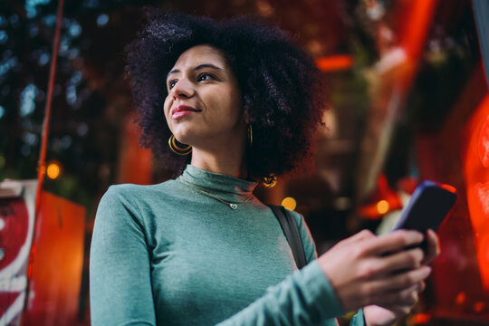 young female user checking smartphone notifications while standing near a bright red neon display window at night, thoughtful Gen Z girl reading instant messages in online chat application