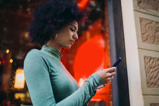 young student typing text messages on mobile smartphone touchscreen under bright red neon storefront glow at night, focused Gen Z youth checking electronic banking app notification