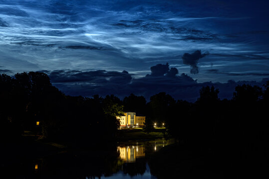 Illuminated manor reflected in a calm river beneath dramatic blue twilight clouds on a peaceful summer night.