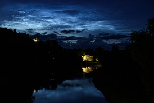 Illuminated manor reflected in a tranquil river beneath glowing noctilucent clouds and a dramatic blue twilight sky.