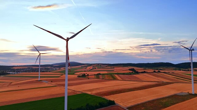 Modern wind generators standing in wide rural fields. High angle shot of alternative energy turbines on brown agricultural lands under twilight sky.