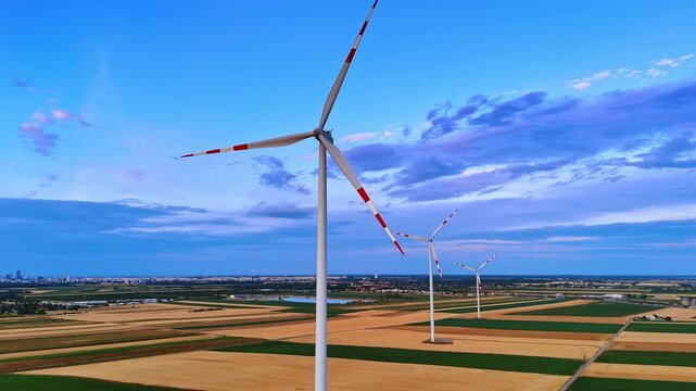 Row of wind turbines across vast agricultural landscape. High perspective shot of ecological generators on geometric fields with faraway urban buildings.