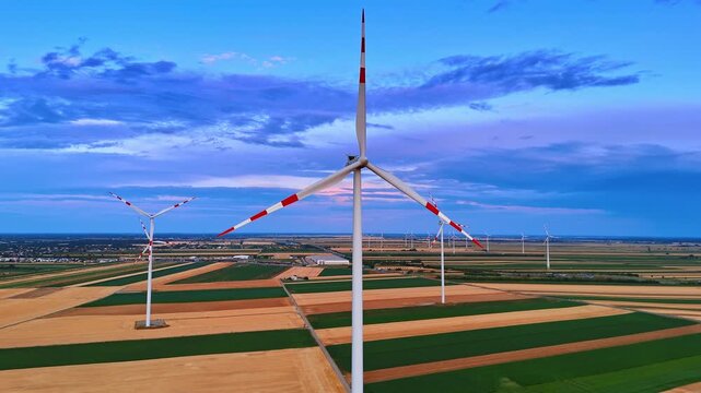 Clean power wind farm over striped agricultural ground. Aerial perspective of ecological turbines on green and brown rectangular land under blue evening atmosphere.