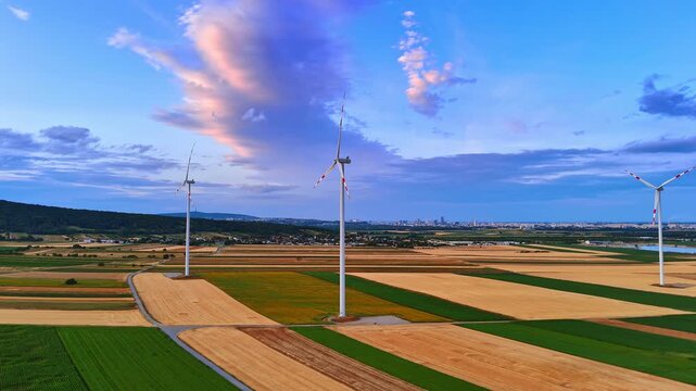 Wind turbines on colorful fields near distant city. Drone view of alternative energy generators over green and golden soil under cloudy sunset sky.
