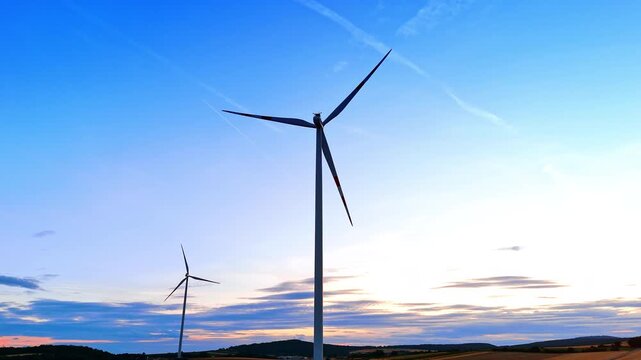 Silhouette of wind turbine under bright evening sky. Upward perspective of industrial propeller with condensation trails on orange twilight background.