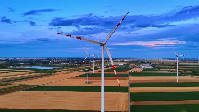 Eco wind generators on dark patchwork fields at dusk. Aerial panorama of multiple electrical turbines over rural land under deep blue sky with orange glow.