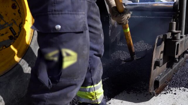 Worker Loading Shovel From Asphalt Paver Hopper Slow Motion. Road construction worker wearing boots and gloves scoops hot asphalt with a shovel near machine hopper wings during street resurfacing.