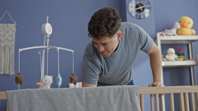 Man leans over crib reaching into cradle and adjusting mobile with blanket on rail and toys on shelf in building; tenderness parenting.