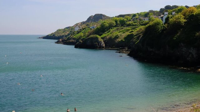 Crystal-clear waters of Balscadden Bay beneath the Howth Cliffs in Ireland. Secluded cove with swimmers, rugged shoreline and emerald hills creating picturesque coastal escape on warm summer afternoon