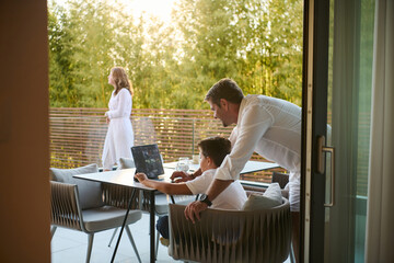 Fototapeta premium Father And Son Working Together On Laptop At Home Balcony While Woman Looks Out Over Terrace