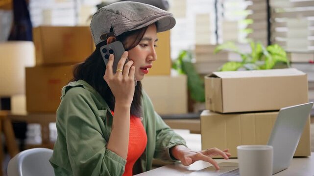 Young Asian female merchant speaking on a mobile phone and looking at a laptop screen. Small business owner giving customer support or confirming delivery details with parcel boxes background.