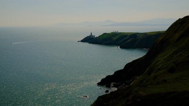 Sweeping vista across dramatic Howth Cliffs featuring Baily Lighthouse, rugged headlands, shimmering Irish Sea, distant mountains, and a passing boat, warm afternoon light, Dublin&rsquo;s scenic peninsula.