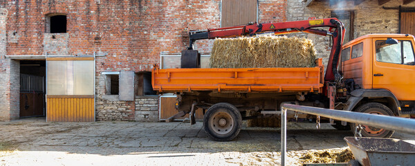 Fototapeta premium Orange farm truck with crane carries hay bales beside brick stable yard, showing rural work and equestrian life. Sunlight, straw, stone walls and barn doors create authentic countryside atmosphere