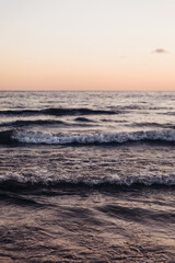 Fototapeta premium Close-up of dark, moody ocean waves rolling and breaking near the shore. Textured water surface and sea foam reflect soft, warm light of twilight, creating a dramatic and abstract marine background.