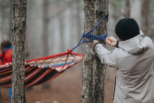 A person ties a blue rope around a tree to secure a striped hammock while a friend waits nearby. Friends in the mountain forest prepare outdoor gear and set up a hammock for relaxation.